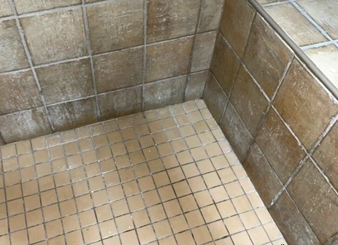 A worn grey tiled hotel shower with stained grout before restoration